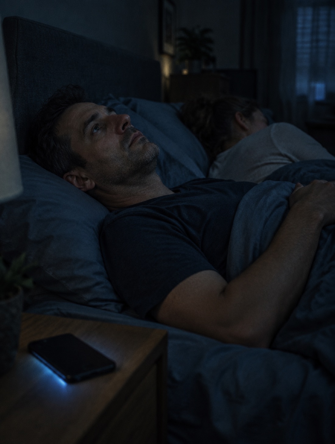 A man lying awake in a dark bedroom at night, his partner asleep beside him.
