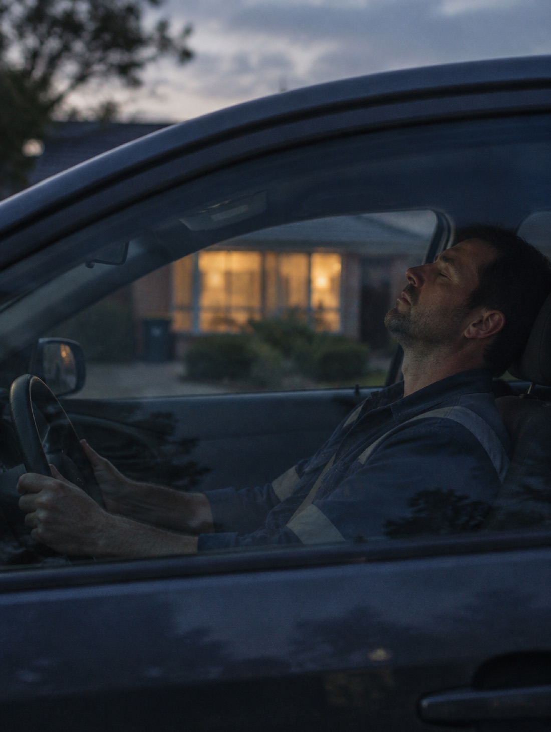 A man sitting in his parked car at dusk with his eyes closed, his house lit behind him.